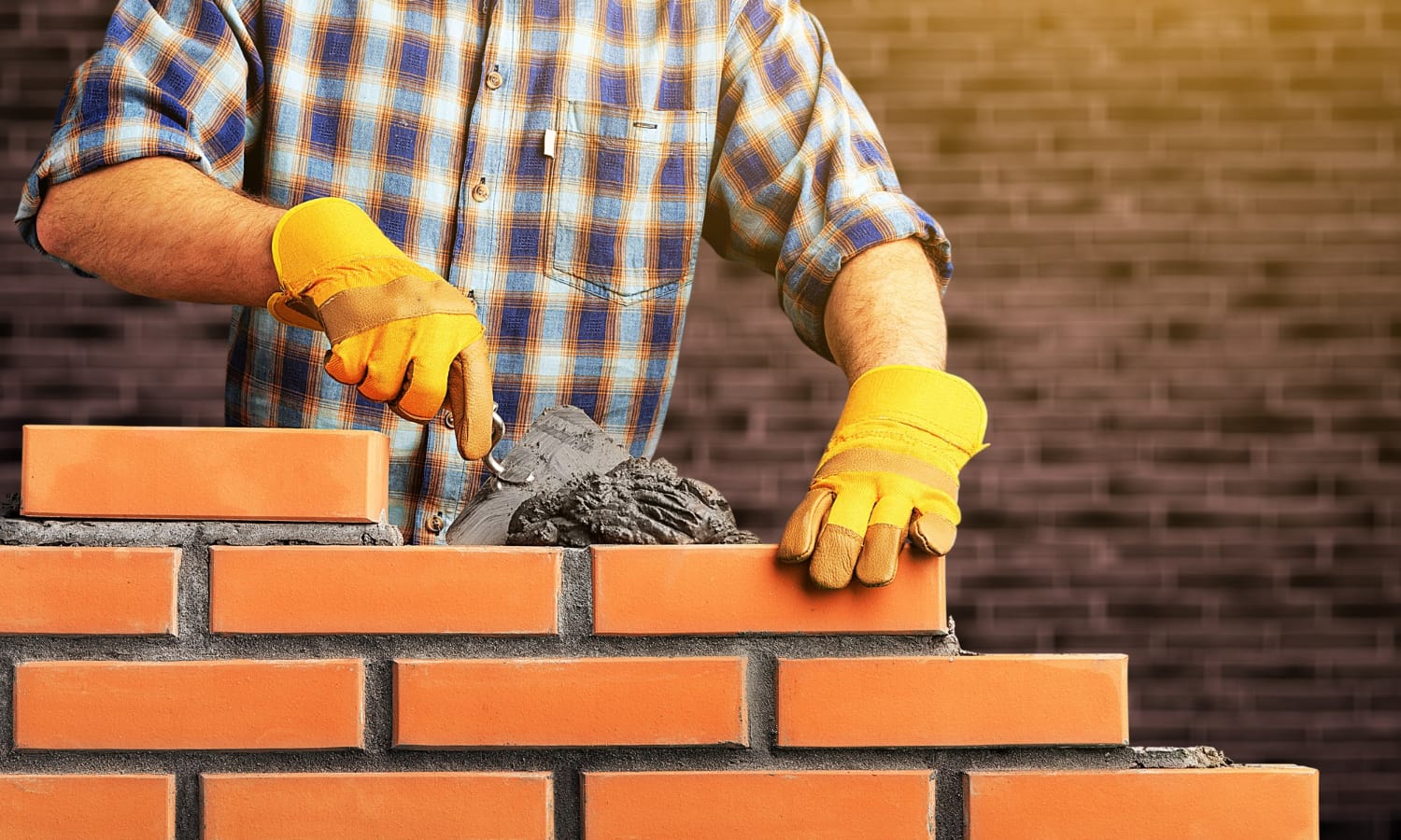 Masonry Contractors Person laying bricks with mortar while wearing yellow gloves, against a blurred brick wall background.