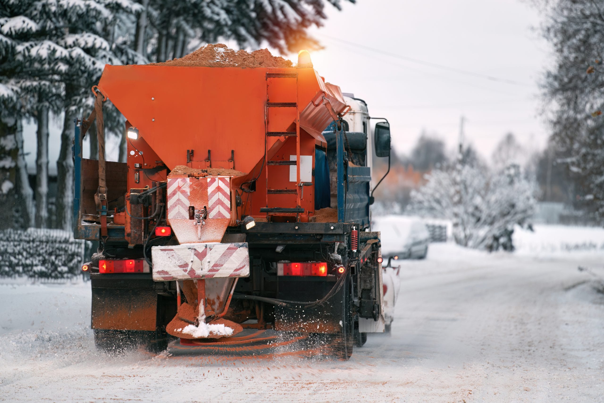 Road maintenance in winter. a tractor equipped with a spreader sprays a salt and sand mixture on the icy and snowy road. The vehicle belongs to the municipal service that melts the snow Snowplow truck spreading salt and sand on a snowy road surrounded by trees.