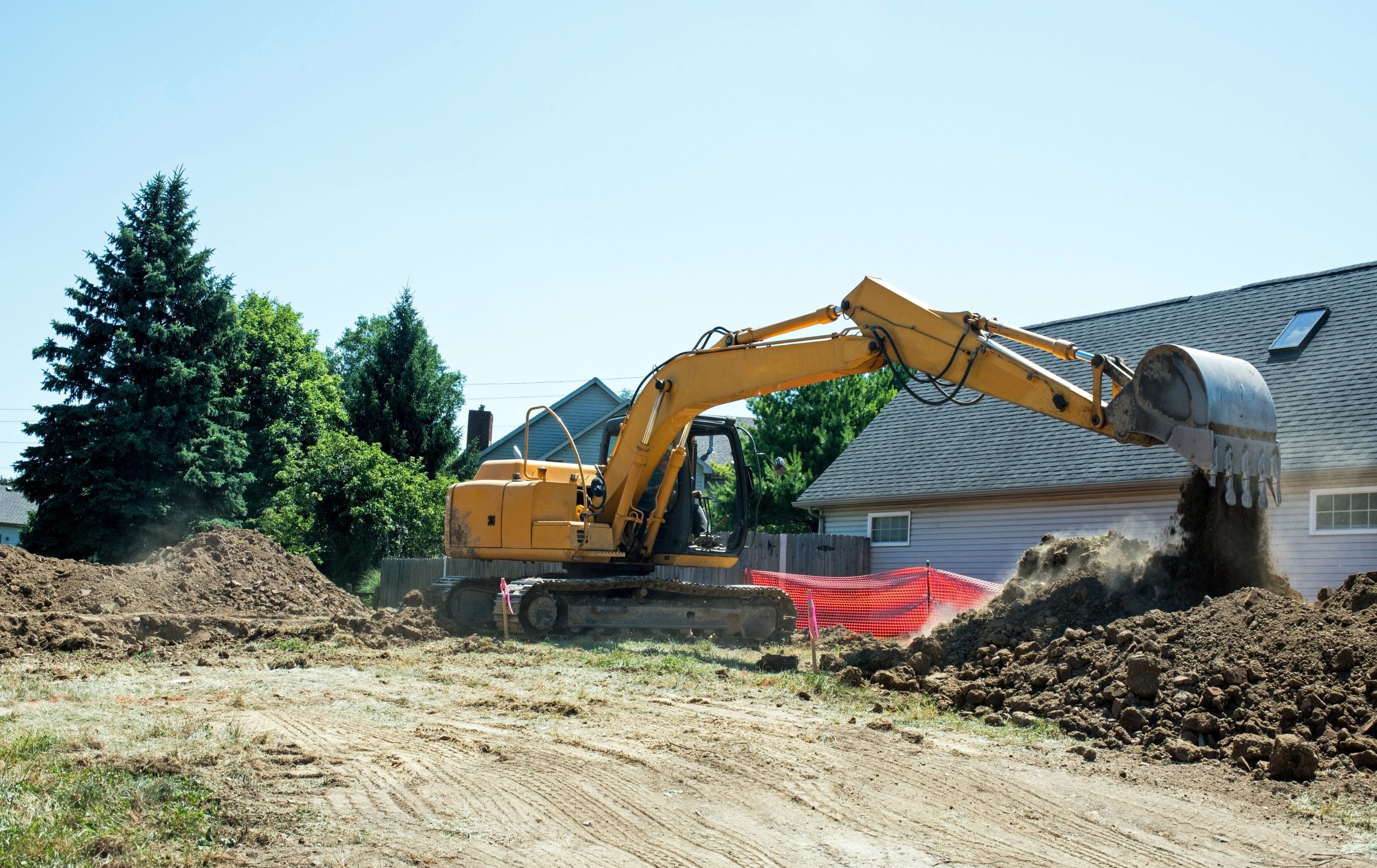 Yellow excavator moving dirt in a residential area, with trees and houses in the background.