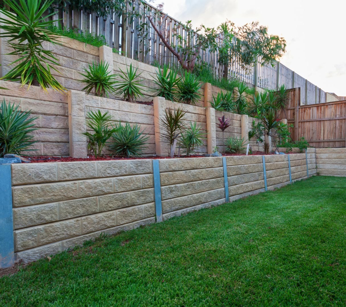 retaining-walls-img-2 Terraced garden with stone retaining walls, green grass, and neatly planted shrubs and small trees.