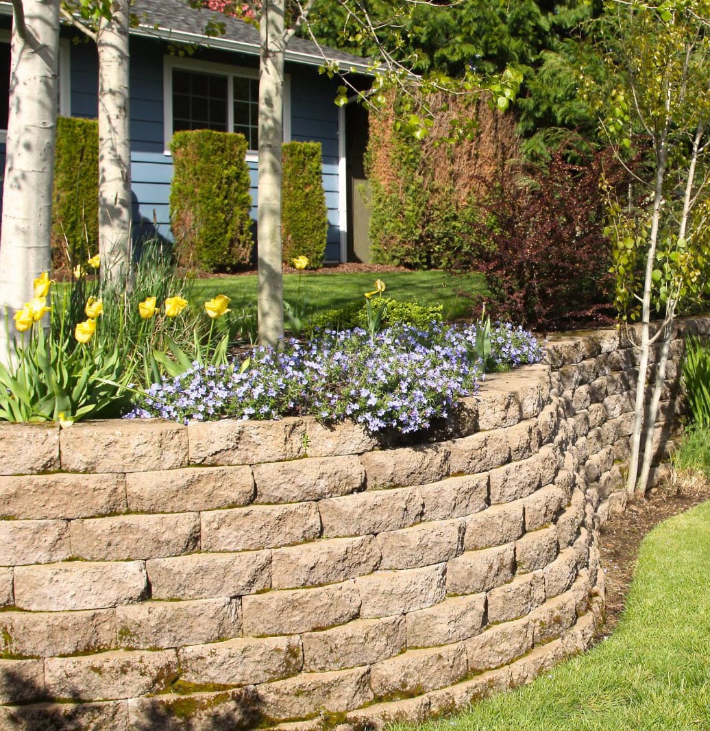 retaining-walls-img A curved stone retaining wall with yellow tulips and purple flowers in front of a house with a blue exterior.