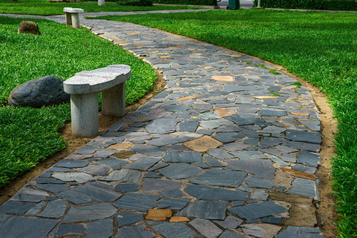 Stone pathway with green grass on both sides and two white stone benches along the path in a park.
