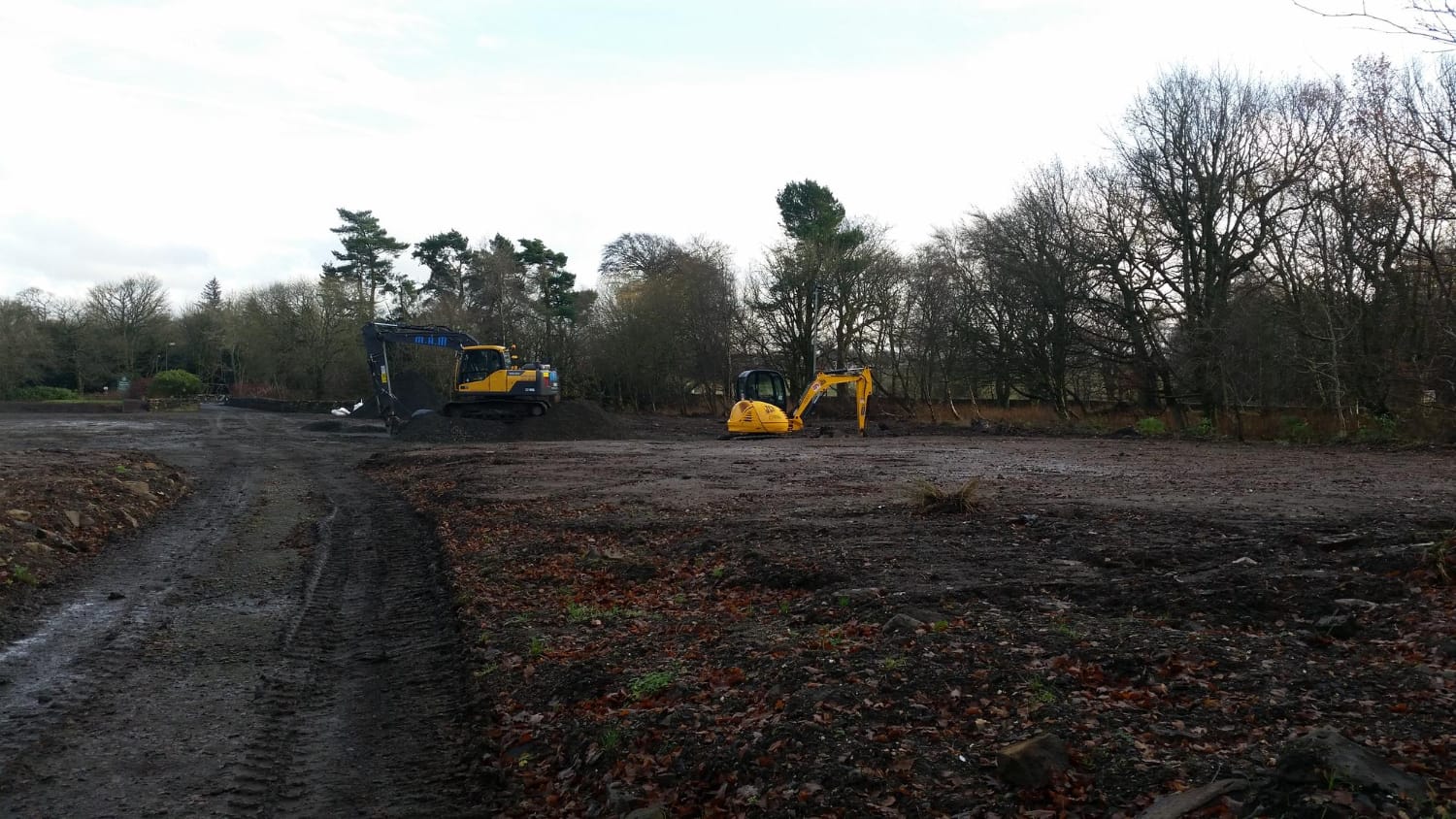 Two yellow excavators on a muddy, cleared construction site surrounded by bare trees and cloudy sky.