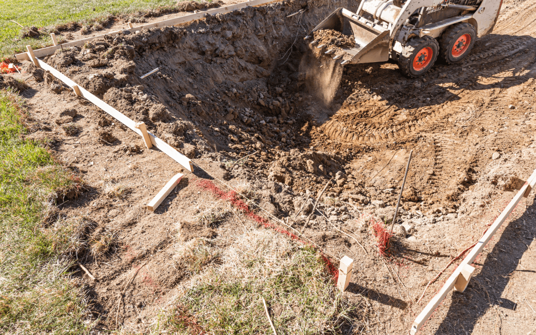 A skid-steer loader dig a foundation in a dirt lot framed with wooden boards and spray paint.