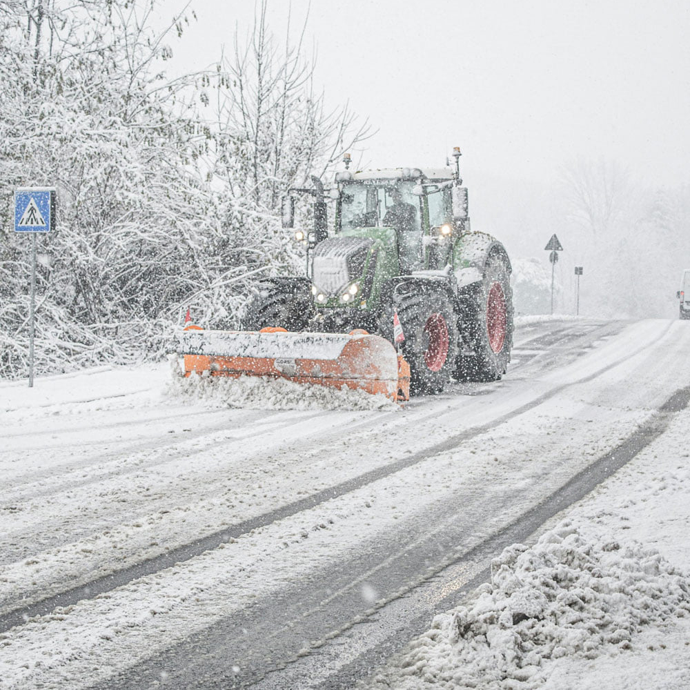 A snowplow clears a snowy road during heavy snowfall in a winter landscape.