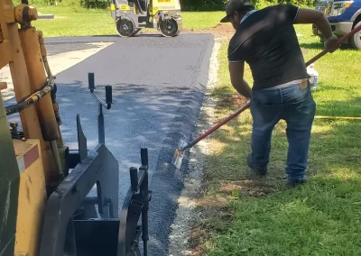 Two workers lay fresh asphalt on a driveway; one smooths the edge while the other operates a roller.