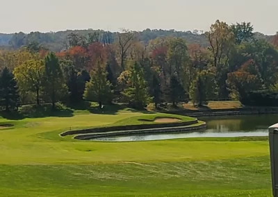 A scenic golf course with green grass, a pond, trees with autumn foliage, and clear sky.