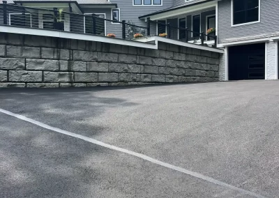 Freshly paved driveway leading up to a modern gray house with large windows and a stone retaining wall.