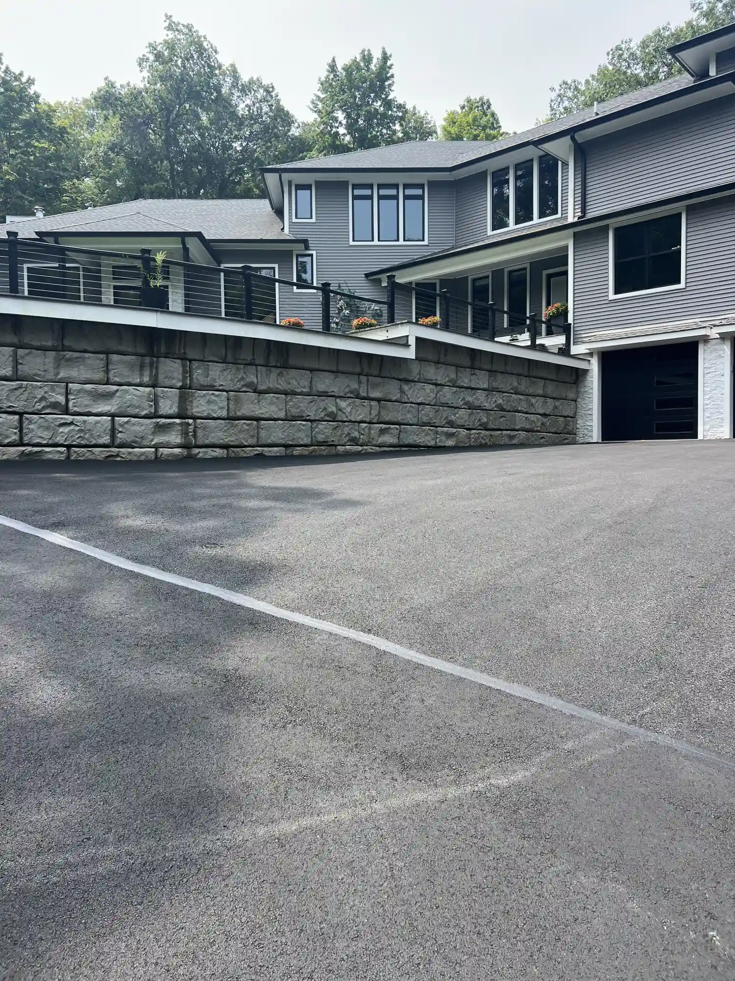 IMG_2404 Freshly paved driveway leading up to a modern gray house with large windows and a stone retaining wall.