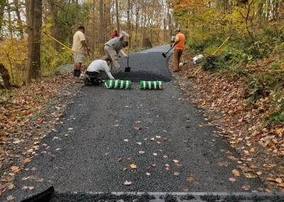 Five people paving a rural road surrounded by autumn trees, with equipment and asphalt rolls visible.