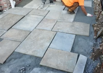 Two workers lay large stone tiles on the ground next to a brick wall at a construction site.