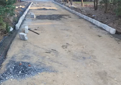 A long dirt driveway under construction, bordered by stone curbs, leads to a house among trees.