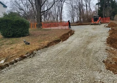 A gravel driveway under construction, with orange fencing and trees in the background on a cloudy day.