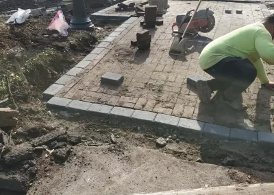 A worker installs bricks on a driveway while construction materials are scattered nearby.