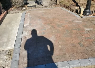 A person works on installing brick pavers, while a shadow of another person appears in the foreground.