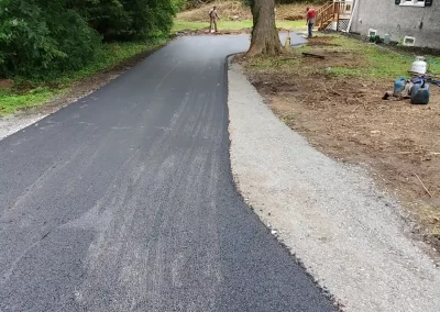 Freshly paved black asphalt driveway curves past a large tree next to a house; people work in the background.