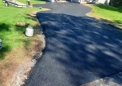 Freshly paved black asphalt driveway in front of a two-story house, surrounded by green grass and trees.