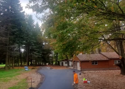 A paved driveway curves past trees and a wooden house; a person in orange stands near the driveway.
