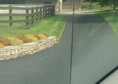 View through a car windshield of a driveway, garden, fence, and a white barn with a traffic cone ahead.