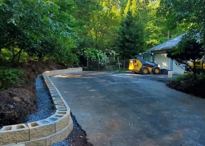 A paved driveway leads to a house, with a retaining wall being built and trees lining both sides.