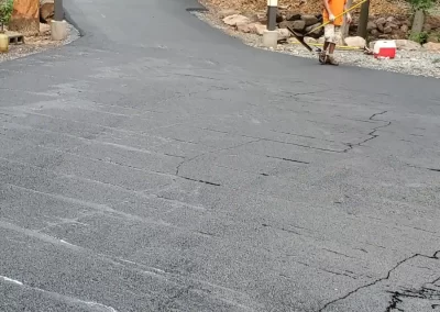 Person in orange safety vest stands by newly paved road near forested area with construction vehicles ahead.