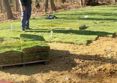 A person stands on newly laid sod in a yard next to a forest, with soil and tools in the foreground.