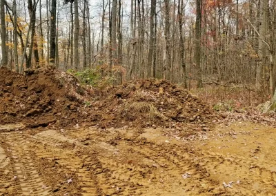 Cleared dirt area with tire tracks, surrounded by piles of soil and autumn trees in a forest.