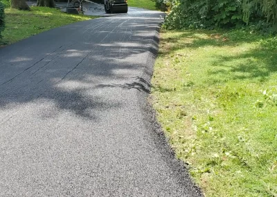 A newly paved road curves through greenery, with a yellow compactor parked in the distance.