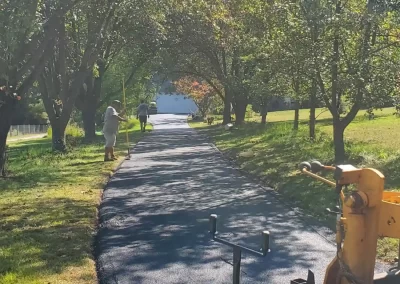 A paved path is being laid under arching trees, with workers and machinery visible on a sunny day.