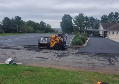 Workers operate a yellow paving machine on a parking lot near a building under a cloudy sky.