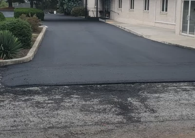 A freshly paved asphalt driveway leading to a beige building with a covered entrance, bordered by shrubs.