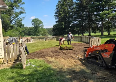 Two people lay sod in a backyard near a wooden fence, with a small orange tractor nearby.