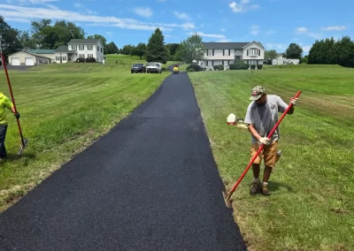 Two workers smooth fresh asphalt on a driveway in a grassy suburban area under a partly cloudy sky.