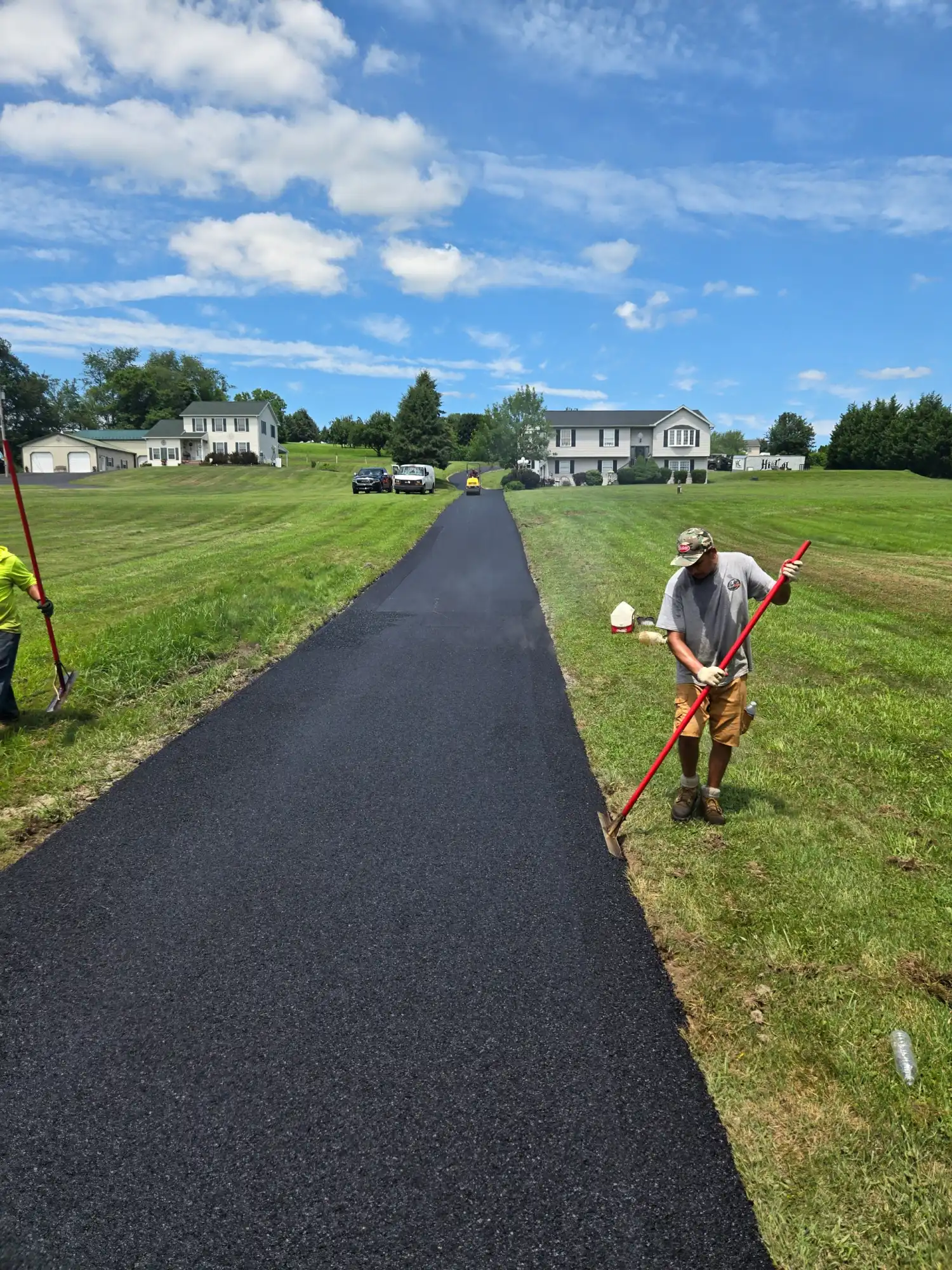 Resized_20250718_133126 Two workers smooth fresh asphalt on a driveway in a grassy suburban area under a partly cloudy sky.