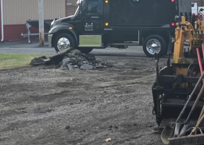 A dark truck and construction equipment at a worksite with broken asphalt and tools in the foreground.