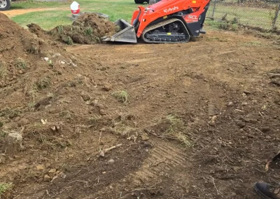 A person operates an orange Kubota skid steer loader, leveling dirt in a yard near a fence and vehicles.