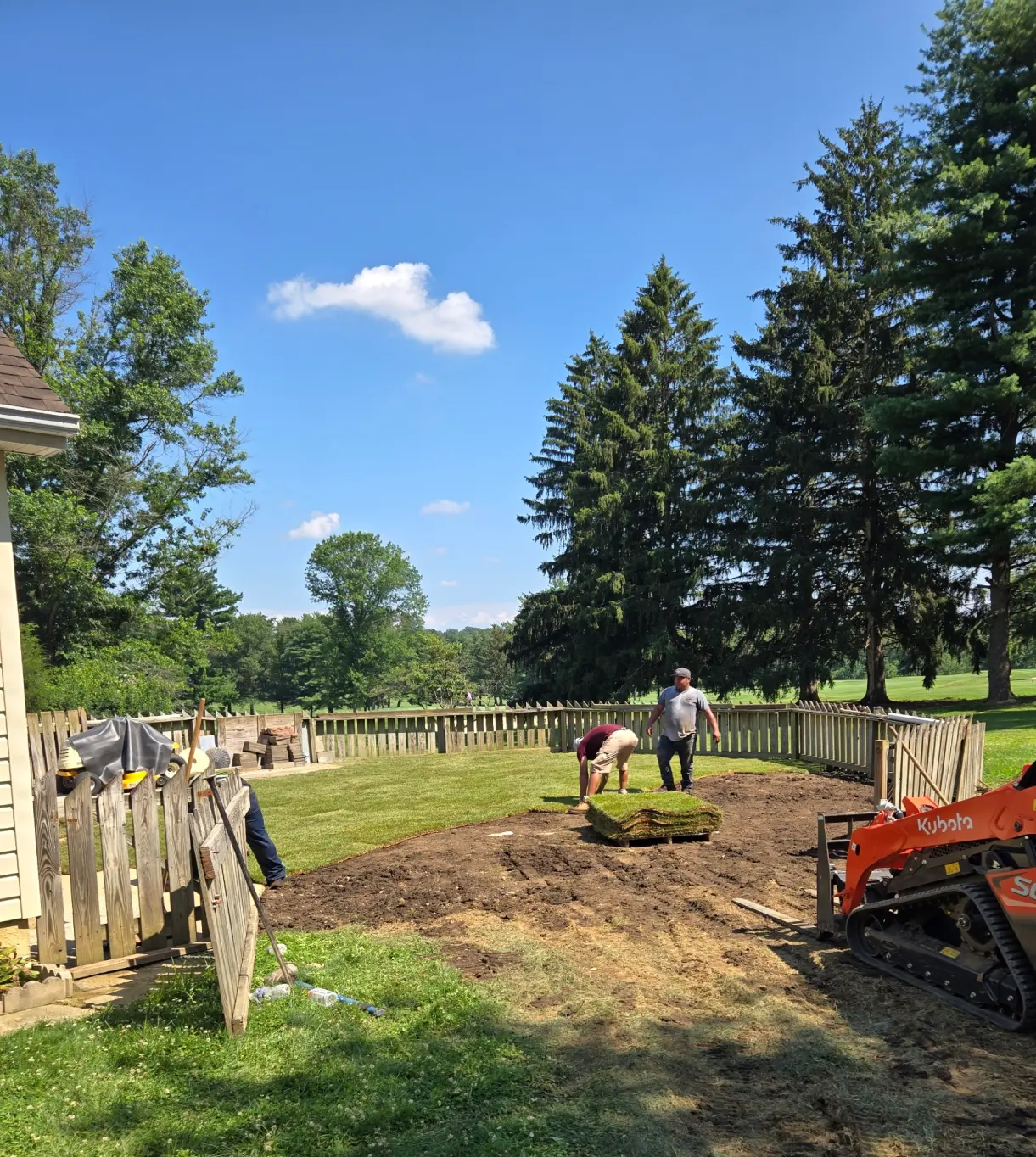Two people lay sod in a fenced yard, with trees and a small orange loader in the background.
