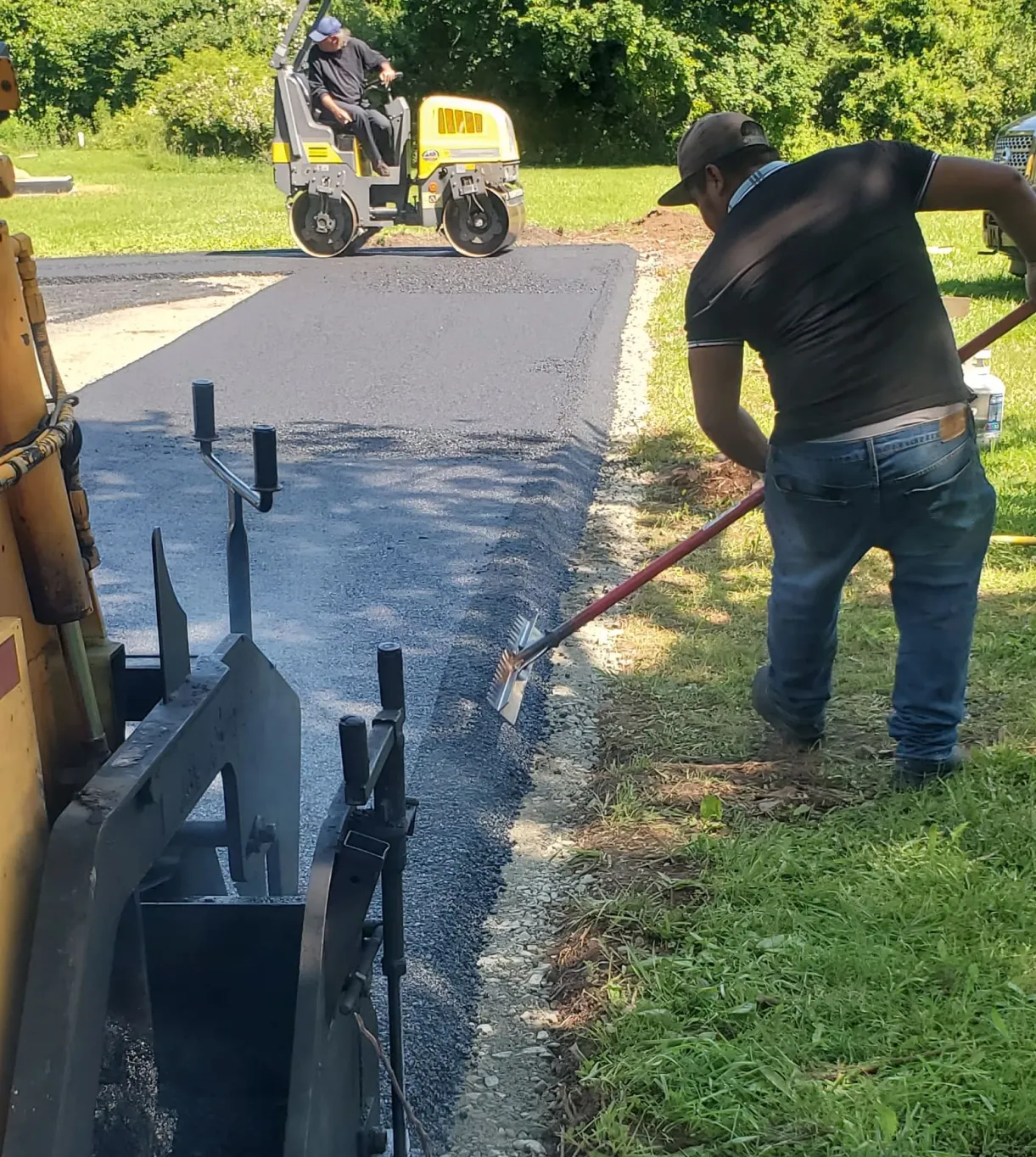 about-2 A worker spreads asphalt while another operates a roller on a newly paved road near grass and trees.