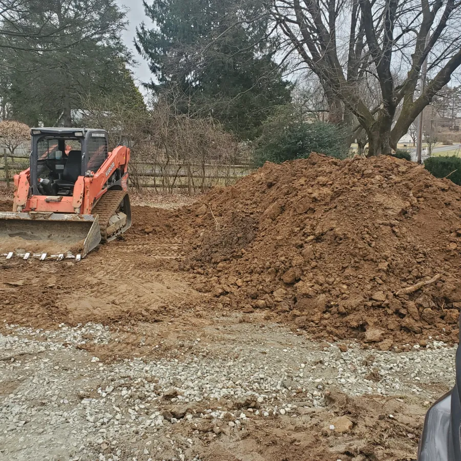 A bulldozer next to a large pile of dirt in a yard with trees and gravel on the ground.
