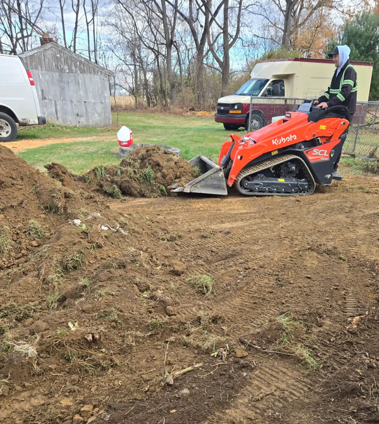 A person operates an orange skid steer loader, moving dirt in a yard with vans and trees in the background.