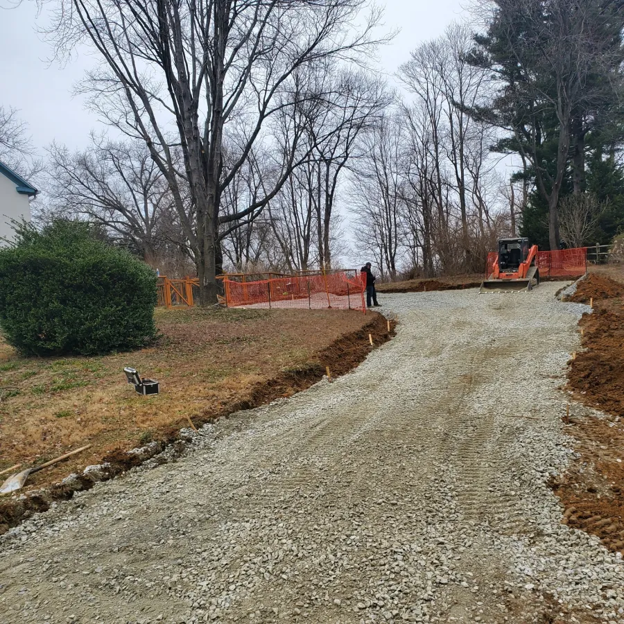 A gravel driveway under construction leads uphill past trees and orange safety fencing, with a worker and machinery.