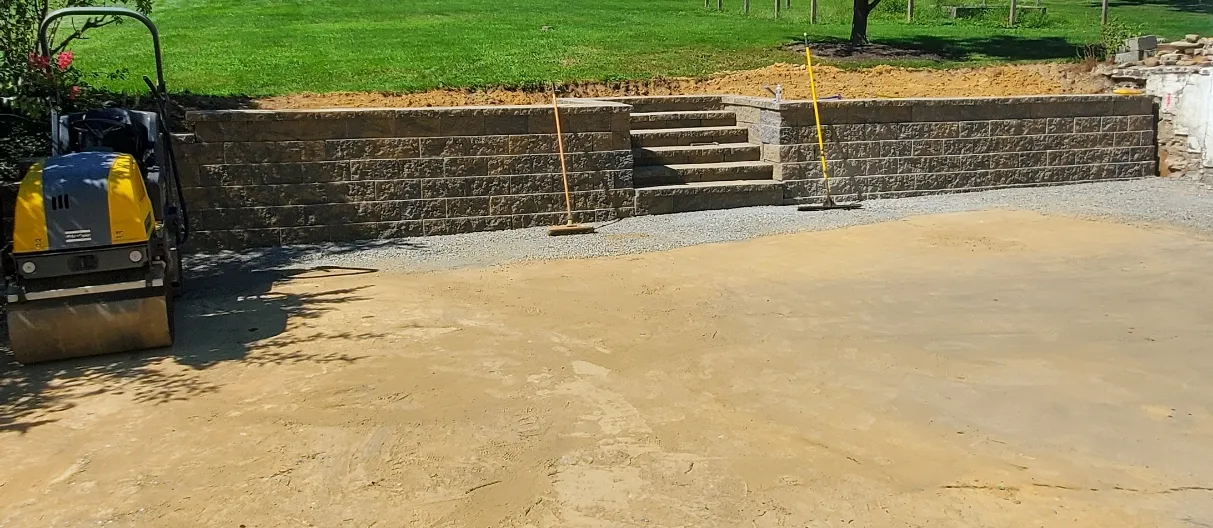 A compact roller and shovels beside a new retaining wall with stairs and fresh gravel on a sunny lawn.