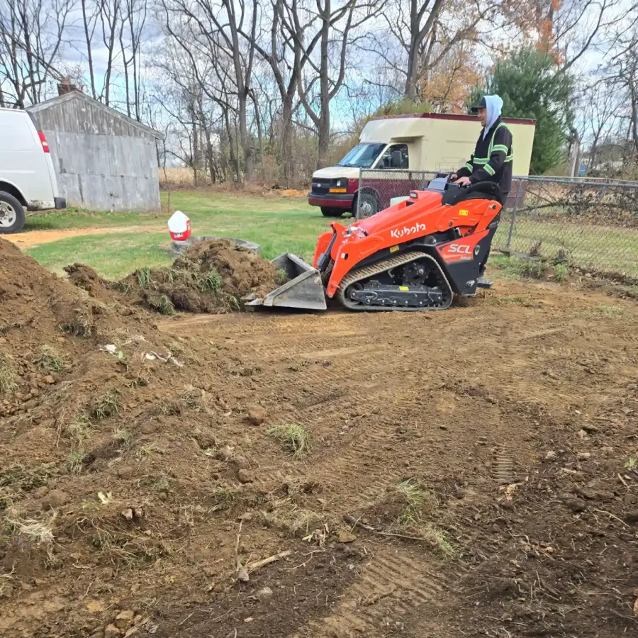 Person operating an orange mini skid steer loader to move soil in a yard next to two parked vans.