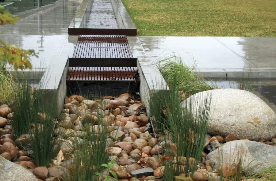 A small metal bridge crosses a rocky stream bed with plants and large stones alongside a concrete path.